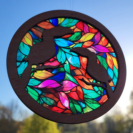 an image of a wooden suncatcher in the shape of a circle. There are 2 toucans laser cut from the top layer. The transparent acrylic background shows a colorful array of leaves