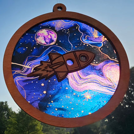 an image of a wooden, circular, suncatcher with a rocket ship laser cut in the center. The transparent acrylic background depicts the solar system with blues and purples