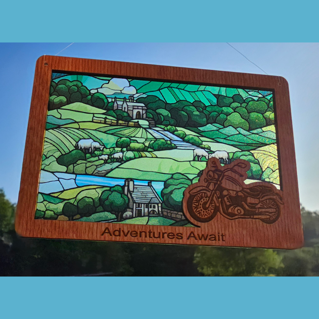 wooden suncatcher in the shape of a rectangle with rounded corners. Has a motorcycle laser etched in the bottom right corner and the text "Adventures Await" along the bottom. It has a transparent acrylic background that depicts a stained glass countryside scene with rolling green hills and spotted with sheep and cottages