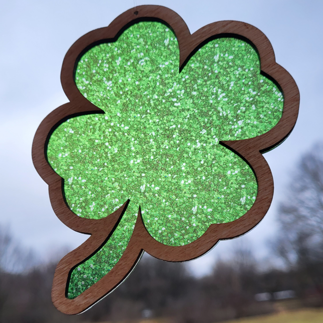 wooden suncatcher in the shape of a four leaf clover. transparent acrylic background with hues of green and specks of gold colors