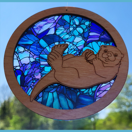 round, wooden suncatcher with a laser etched image of a sea otter holding an oyster shell. Has a transparent acrylic background of various seashells in hues of blue and purple