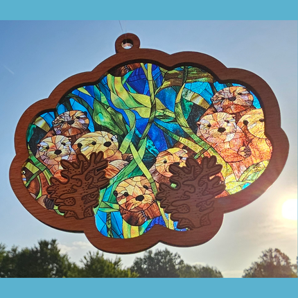 wooden suncatcher in the shape of an oyster shell with batches of seaweed laser etched near the bottom. Transparent acrylic background of a family of sea otters peeking out from the seaweed. The background depicts a stained glass look in its detail.