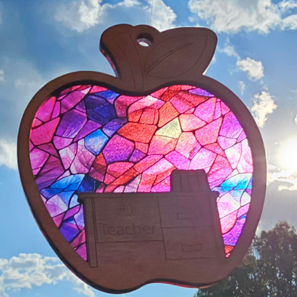 wooden suncatcher in the shape of an apple with a desk laser engraved into the lower portion with the words #1 Teacher. The transparent acrylic background shows a variety of pink, red, and purple colors. Teacher name can be personalized