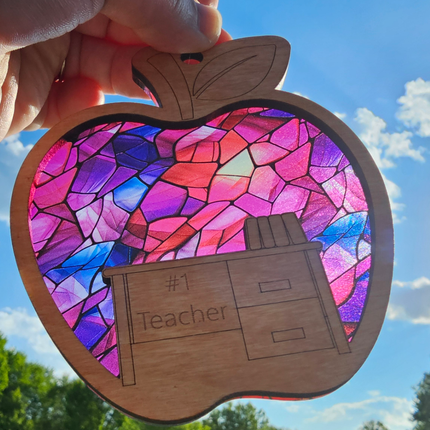 wooden suncatcher in the shape of an apple with a desk laser engraved into the lower portion with the words #1 Teacher. The transparent acrylic background shows a variety of pink, red, and purple colors. Teacher name can be personalized