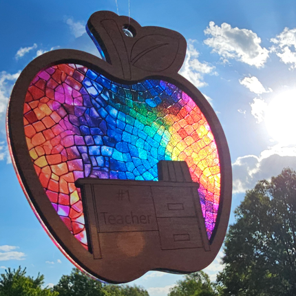 wooden suncatcher in the shape of an apple with a desk laser engraved into the lower portion with the words #1 Teacher. The transparent acrylic background shows a variety of rainbow colors.