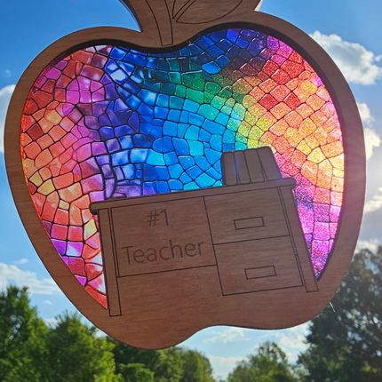 wooden suncatcher in the shape of an apple with a desk laser engraved into the lower portion with the words #1 Teacher. The transparent acrylic background shows a variety of rainbow colors. Teacher name can be personalized